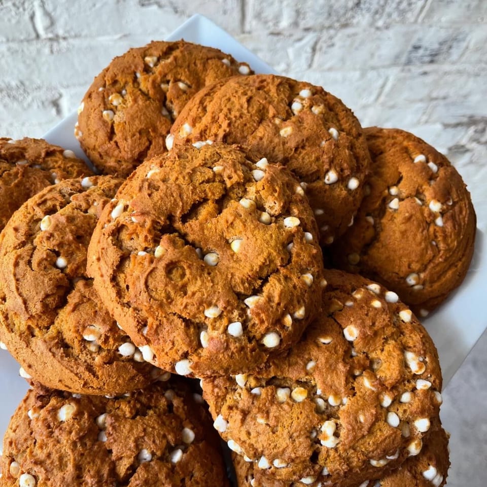 A white plate piled high with round, golden-brown vegan cookies studded with white chocolate chips.