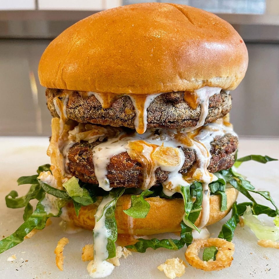 A double-stacked vegan burger with two patties, white sauce, orange sauce, and lettuce on a bun.