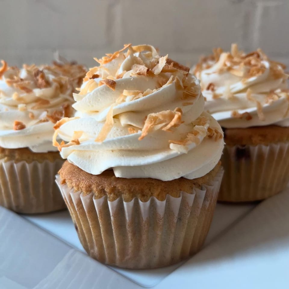 Three vegan cupcakes with white frosting and toasted coconut flakes on a white plate.