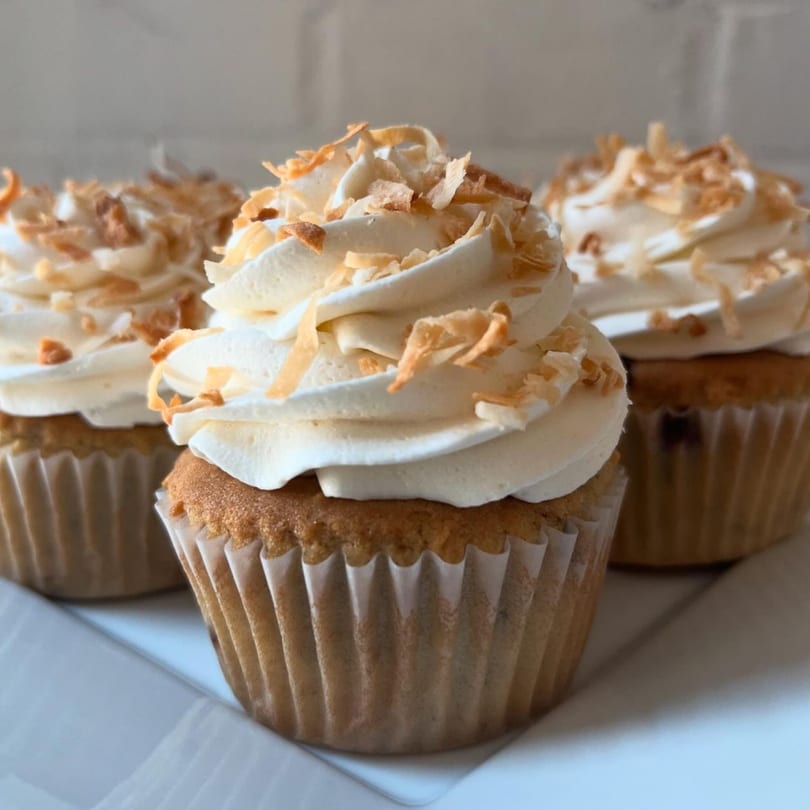 Three vegan cupcakes with white frosting and toasted coconut flakes on a white plate.