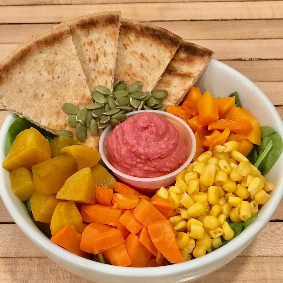 A colorful vegan bowl with pita bread, pink hummus, pumpkin seeds, corn, carrots, and squash.