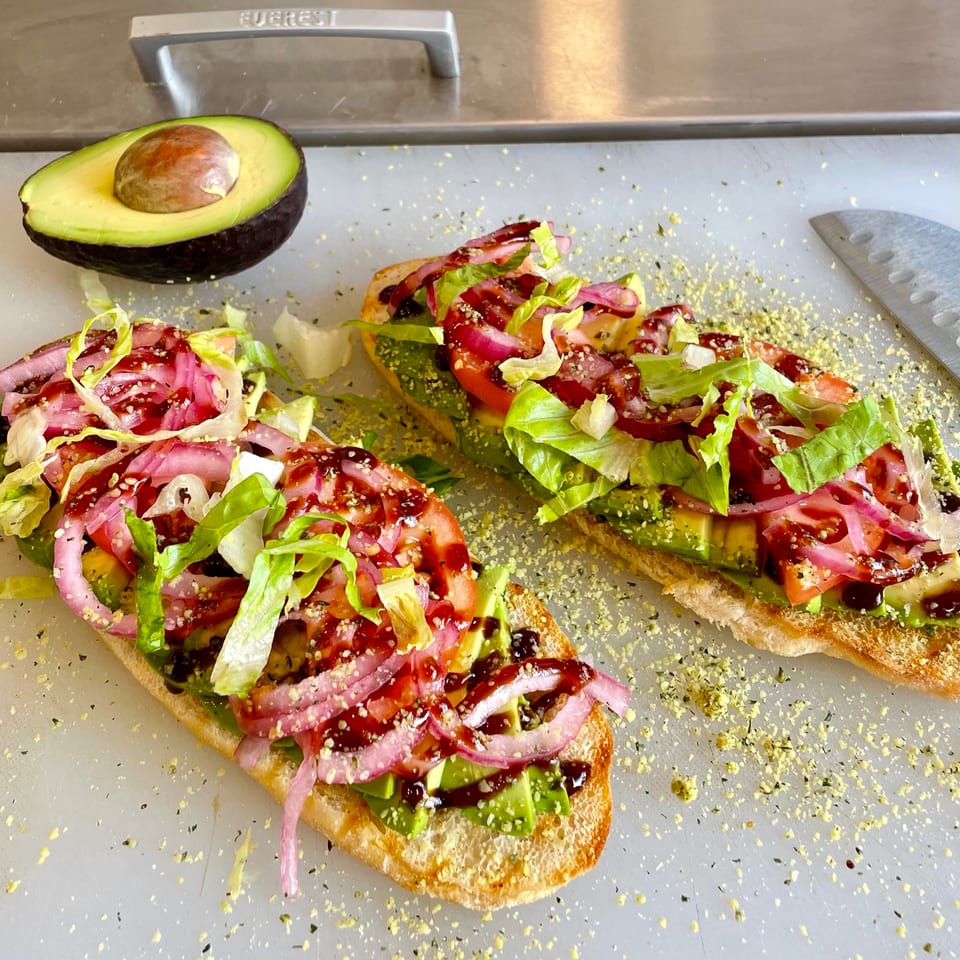 Two vegan avocado toasts with lettuce, tomato, pickled onions, and balsamic glaze, next to a halved avocado.