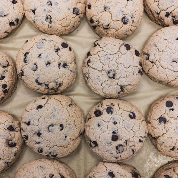 Close-up of several vegan chocolate chip cookies with cracked tops on a light brown surface.