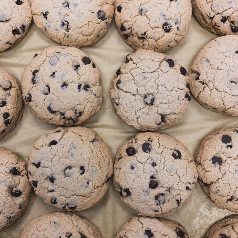 Close-up of several vegan chocolate chip cookies with cracked tops on a light brown surface.