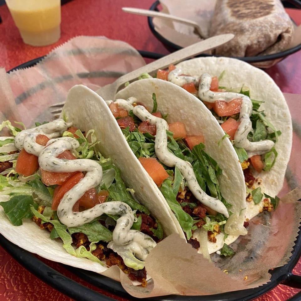 Three vegan tacos with lettuce, diced tomatoes, and creamy sauce, served with a drink and burrito.